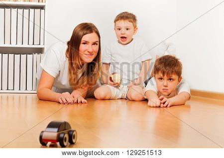 Mother and her two sons playing with toy wooden car laying at the floor