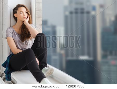 people, emotion and teens concept - sad unhappy pretty teenage girl sitting on windowsill and looking through window over singapore city skyscrapers background