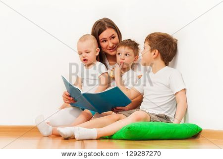 Young mother reading book to her three age-diverse kids, sitting on green pillow against white wall at the room