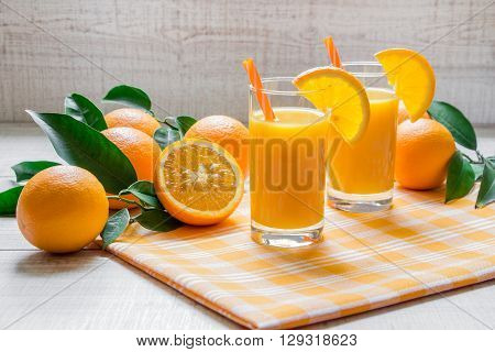 Right 2 glasses of orange juice on yellow checkered napkin left and around freshly picked oranges with green leaves on light wooden background. Horizontal. Daylight.