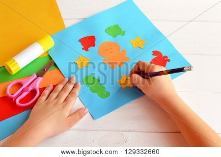 Child holds a pencil and draws. Child doing a card with sea animals and fish. Sheets of colored paper, scissors, glue, set for kids art