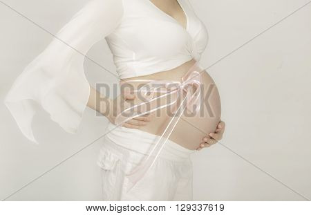 Torso of a pregnannt woman with pink ribbin on her abdomen dressed in white on gray background.