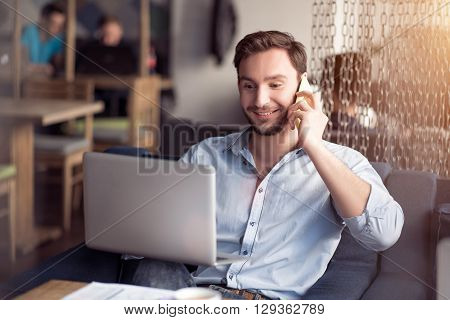 Lively conversation. Cheerful positive content bearded man using laptop and sitting at the table while  talking on cell phone