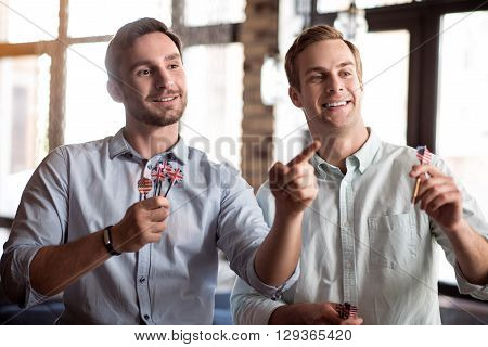 Have some entertainment. Positive handsome delighted smiling friends playing darts and expressing joy while spending time together