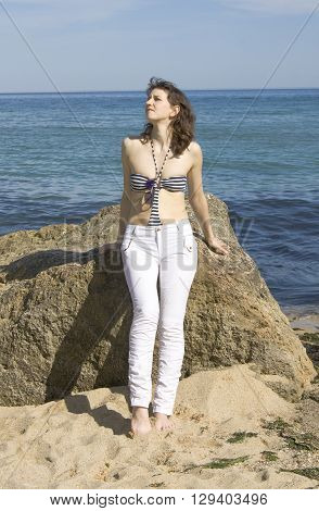 Young European woman with brown hair in white trouses and top of bikini sits on big stone on beach.