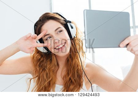 Happy woman in headphones making selfie photo on tablet computer at home