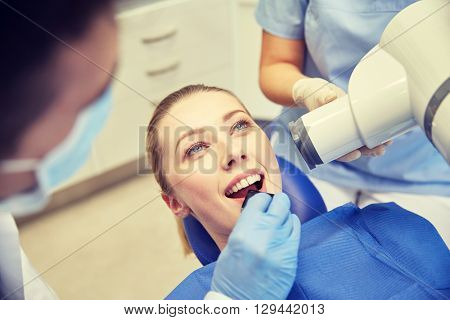 people, medicine, stomatology, technology and health care concept - close up of dentist putting intraoral shield to female patient mouth and assistant directing x-ray machine at dental clinic