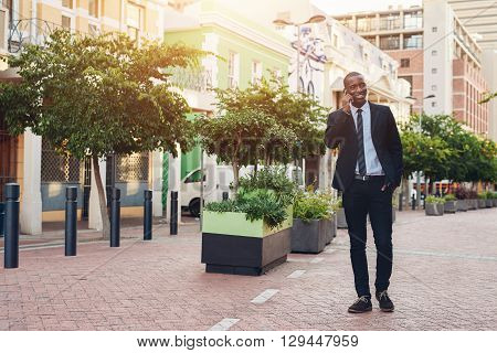 Full length shot of a handsome young African businessman in a contemporary suit walking casually along a city sidewalk with plants, smiling confidently while talking on his mobile phone