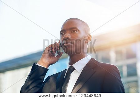 Young African businessman holding his phone to his ear and looking seriously into the distance with buildings and sunflare behind