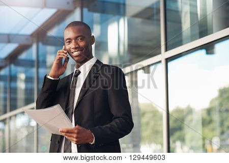 Young African businessman standing outside in front of the reflective windows of a big city building holding a file, and his phone to his ear while smiling at the camera