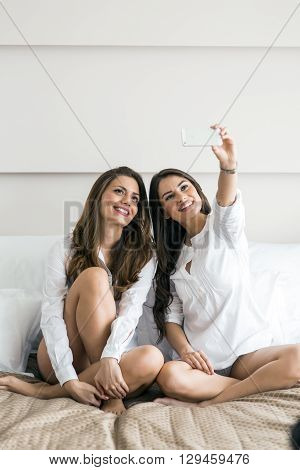 Two beautiful women lying on a bed taking a photo of themselves with a phone in a luxorious bedroom