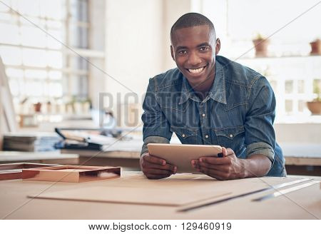Smiling African small business owner, leaning on a work table in his studio, smiling at the camera while holding a digital tablet