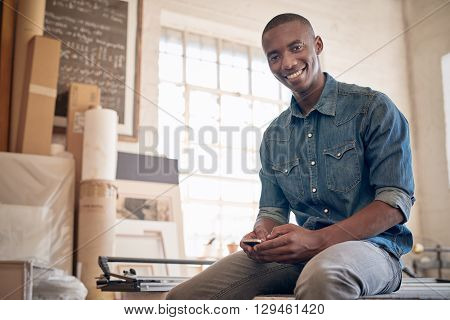 Low angle portrait of a handsome young business owner of African descent, smiling at the camera while holding is mobile phone and sitting on a workbench in his studio workshop