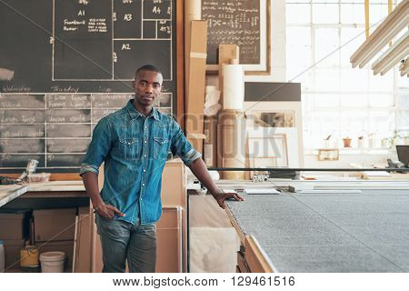 Serious young designer of African descent looking at the camera while standing casually in his studio workshop