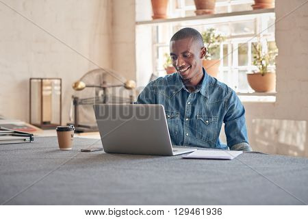 Young designer of African descent, in his design studio workshop with beautiful lighting, smiling while working on his laptop