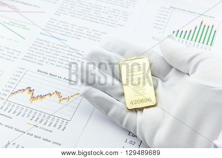 Man right hand with a white handy glove is showing / presenting an one ounce gold bar to a customer for inspection. A gold ingot in a hand on a table with many financial and business analysis reports.