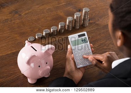 Close-up Of A Businesswoman Using Calculator With Piggybank And Stack Of Coins On Desk
