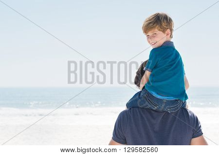 Young boy sitting on father shoulder. Happy son enjoying shoulder ride at beach, copy space. Son looking at camera and smiling at beach.