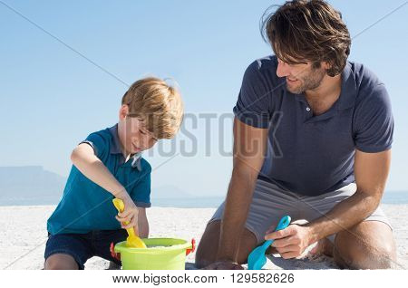 Cute little boy playing with sand at beach. Smiling father and son making sand castles in a summer day. Happy boy filling basket with sand. 