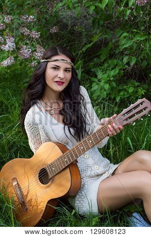 Beautiful hippie girl with guitar sitting on bright grass near lilak bush