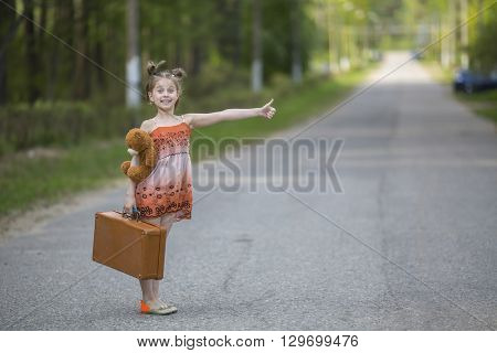 Funny little girl hitchhiker with a suitcase and a teddy bear.