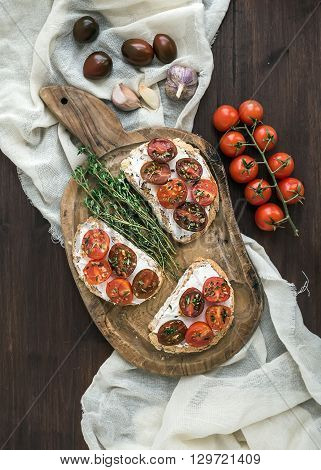 Sandwiches brushtta with roasted cherry tomatoes, soft cheese, garlic and herbs on a rustic wooden board and white kitchen towel over a dark wood background. Top view