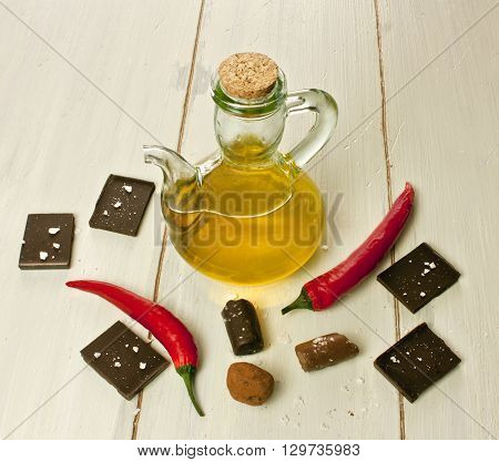 Chocolate pieces and bonbons with unusual chocolate components: sea salt flakes red hot chilli peppers and olive oil in a pitcher on a light wooden background texture with copyspace