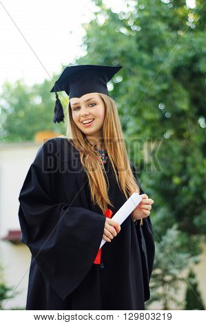 The girl in the mantle keeps in hands graduate diploma and looks into the camera. On her face a smile