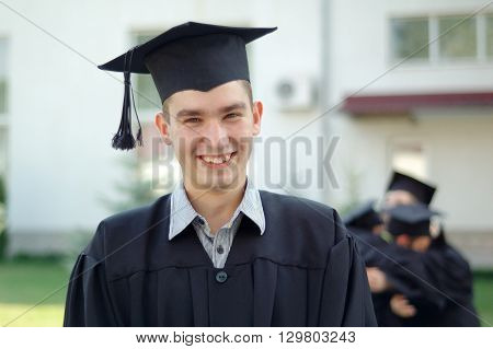 A young Graduate in black mantle with a smile looking at the camera. He has an academic cap on his head.