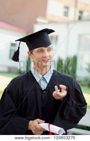 The graduate of the University of something said. He is dressed in a black cloak and a hat. Student holding his diploma.