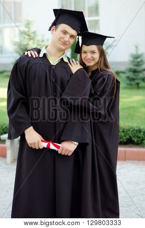 Guy and girl in black robes embracing. In the hands of Man diploma. She put her hands on his shoulders.
