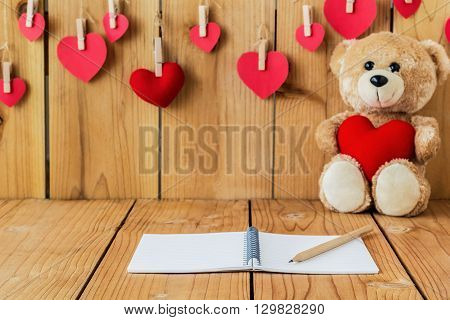 A photo of notepad on wood board with teddy bear holding heart sharp pillow background Selective focus