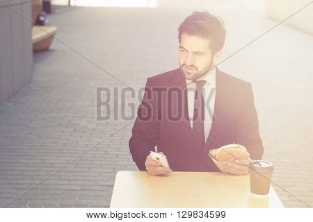 Toned picture of young confident businessman eating and drinkig in cafe. Handsome man sitting at table and looking away.
