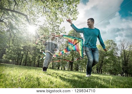 Runnings little boy and father flies with them kite in the park under sunligt.