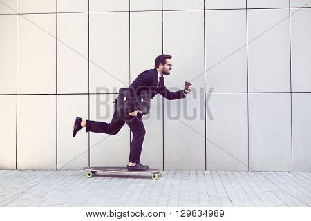 Toned image of businessman in black businesssuit on longboard walking with cup of coffee in his hand. Freelance man hurrying to his office.