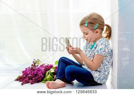 4 years old girl sitting on a windowsill and looking in a small mirror. By lies bouquet of lilacs