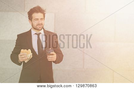 Toned picture of young businessman eating junk food and drinkig tea or coffee while walking to office along street.