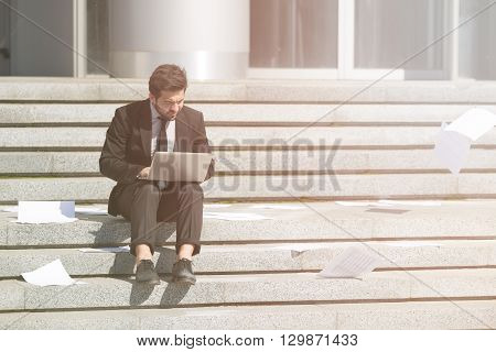 Toned picture of handsome businessman sitting on stairs incity centre and using laptop computer for business purposes.