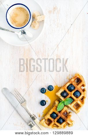 Espresso coffee cup and soft belgian waffles with fresh blueberries and marple syrup on white painted wooden board over light blue background. Top view, copy space