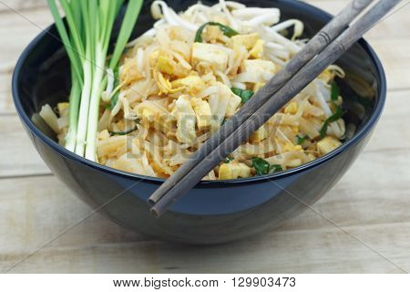 Thai fried noodle in black bowl with bean sprouts and Garlic chives on topping and chopsticks on the bowl. Pad Thai in a black bowl put on wood table.