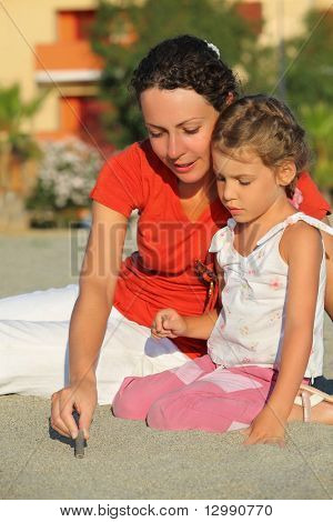 Мать сидит на берегу с дочерью и показывает его как рисовать stick на песке