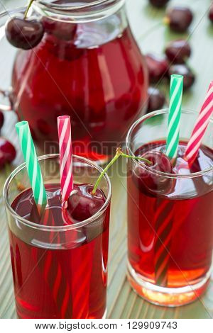 Two glasses of cherry juice with straws in the foreground and in the background a jar full of cherry juice. 2 cherry juices. Vertical. Close-up. Daylight.