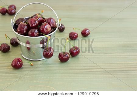 In the frame on the left a small bucket with cherries, around scattered cherries, on the right empty space on a light green background. Bucket of cherries. Horizontal. Daylight.