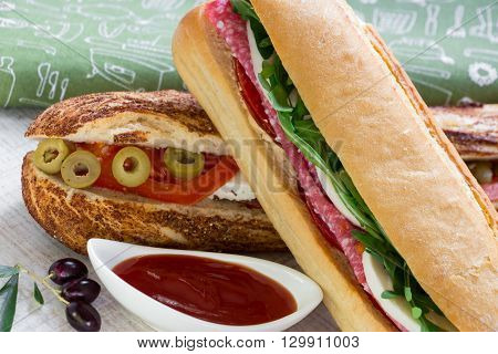 Diagonally lying sandwich with salami, mozzarella, arugula and tomato on sandwich with feta cheese, olives, tomato, near bowl with ketchup, a few olives on the background a green napkin.  Horizontal. Daylight.