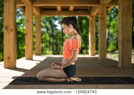 Attractive girl sits sideways on her heels on the black yoga mat on the wooden terrace on the nature background. She holds her hands on her legs. She looks in front of herself with omitted chin. She wears black shorts and orange t-shirt. Outdoors. Horizon