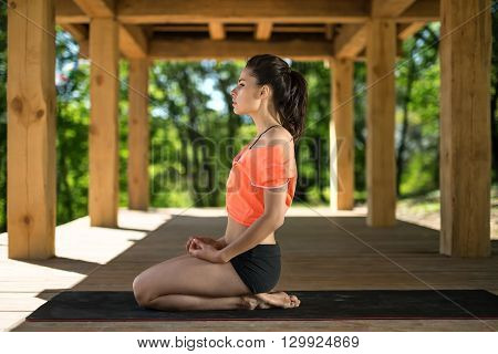 Barefoot girl sits sideways on her heels on the black yoga mat on the wooden terrace on the nature background. She holds her hands on her legs. She looks in front of herself with raised chin. She wears black shorts and orange t-shirt. Outdoors. Horizontal