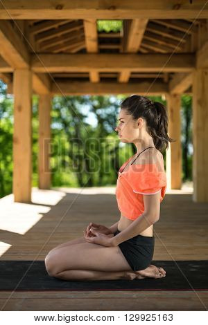 Brunette girl sits sideways on her heels on the black yoga mat on the wooden terrace on the nature background. She holds her hands on her legs. She looks in front of herself. She wears black shorts and orange t-shirt. Outdoors. Vertical.
