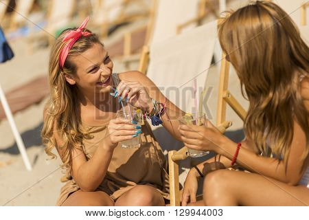 Two beautiful young girls lying on a sunbed on the beach cheering with a lemonade and chatting