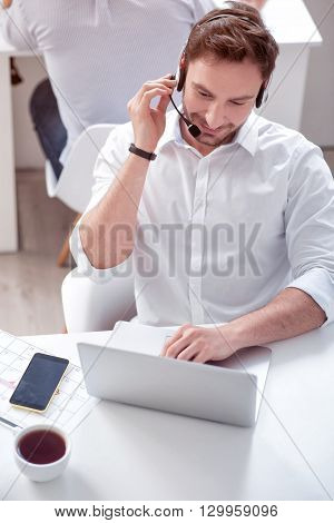 Zealous worker. Positive handsome smiling man sitting at the table and working on the laptop in the office with his colleague while  using headset with micro