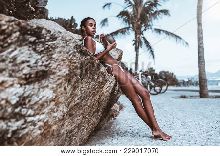Haughty Young Sexy Brazilian Female Is Drinking Fresh Coconut Water While Leaning On The Crag With B